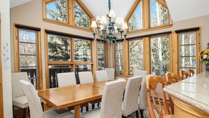 A dining area with a wooden table and chairs, large windows, and a chandelier. - Haymeadow Townhome A1 | 5BR Ski-Out, Private Hot Tub (Avon)