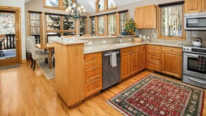 A spacious kitchen with wooden cabinets, a central island, and a large window overlooking a deck. - Haymeadow Townhome A1 | 5BR Ski-Out, Private Hot Tub (Avon)