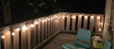 A wooden deck with string lights, two green chairs, and a small table with a floral tablecloth.