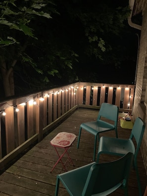 Una terraza de madera con luces colgantes, dos sillas verdes y una pequeña mesa con mantel floreado.