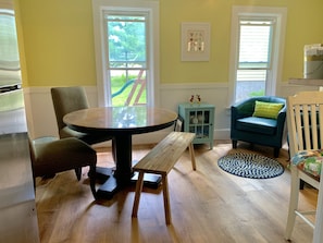 Una habitación con una mesa redonda de madera, dos sillas, un mueble bajo, un sillón azul con un cojín verde y una alfombra estampada.