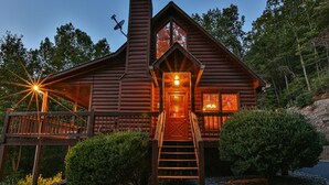 A log cabin with a wooden deck, a central staircase, and a lit interior.