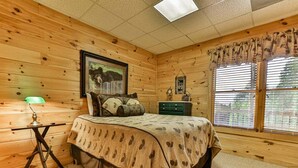 A wooden cabin bedroom with a bed, a desk, a lamp, and a window with blinds.