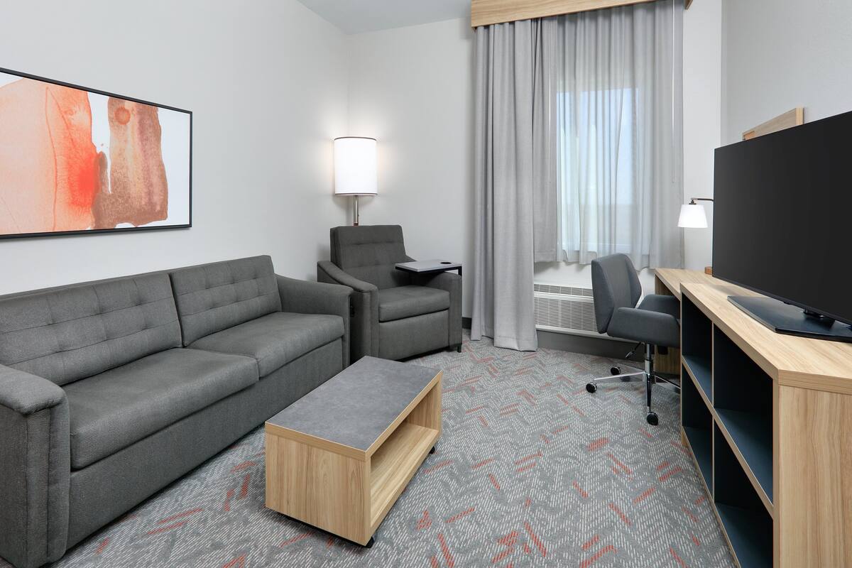 A modern hotel room with a grey sofa, a wooden coffee table, a desk with a flat-screen TV, and a large window with curtains.