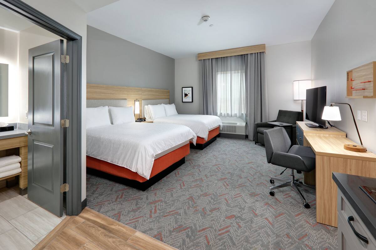 A hotel room with two beds, a desk, a chair, a television, and a window with curtains.