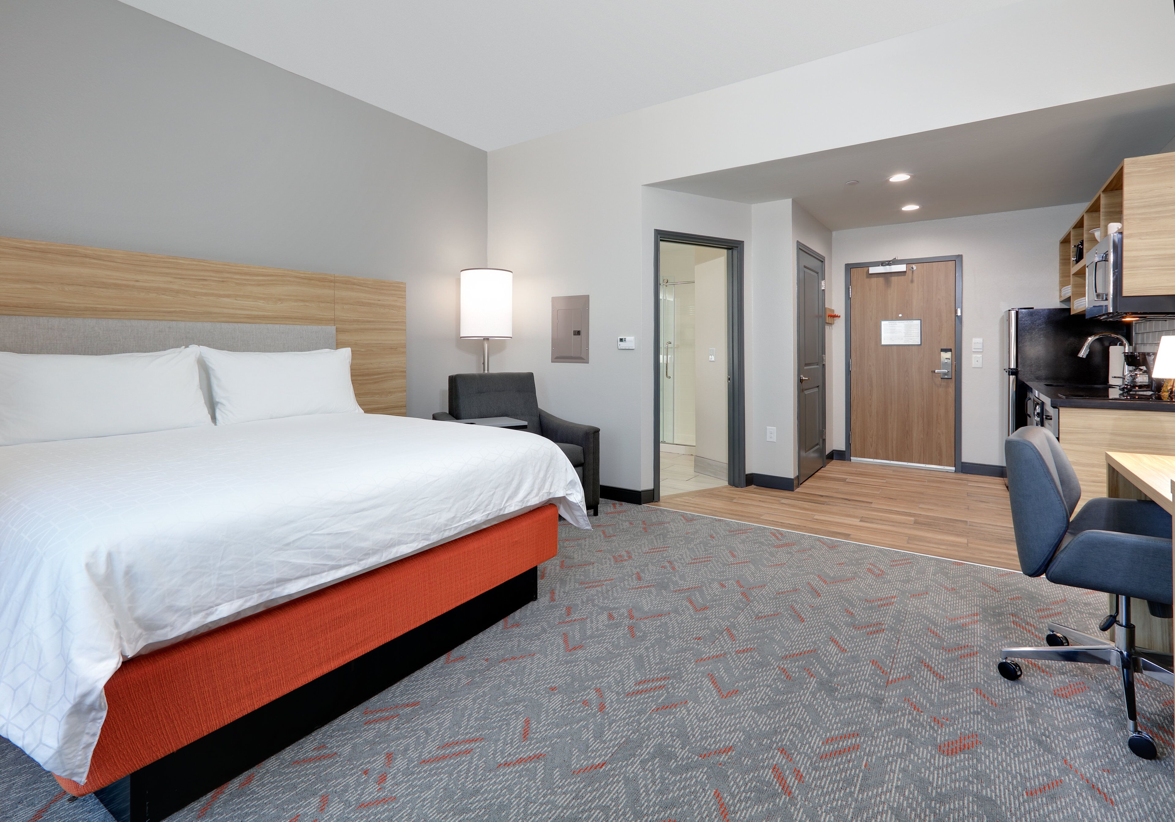A hotel room with a bed, a desk, a chair, a lamp, and a kitchenette.