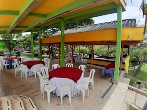 An outdoor covered patio area with round tables and white plastic chairs, surrounded by a green and yellow structure. - Ecohotel La Casona (Pereira)