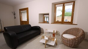 A living room with a black leather sofa, a glass coffee table, a wicker chair, and two windows with wooden frames.