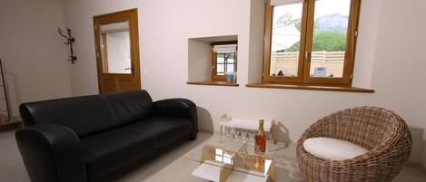 A living room with a black leather sofa, a glass coffee table, a wicker chair, and two windows with wooden frames.
