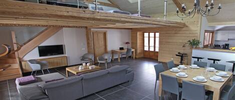 A spacious living area with a high ceiling, wooden beams, a dining table set for six, and a modern kitchen in the background.