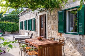 Un edificio de piedra con contraventanas verdes, una mesa de madera con sillas y un cuenco de fruta.
