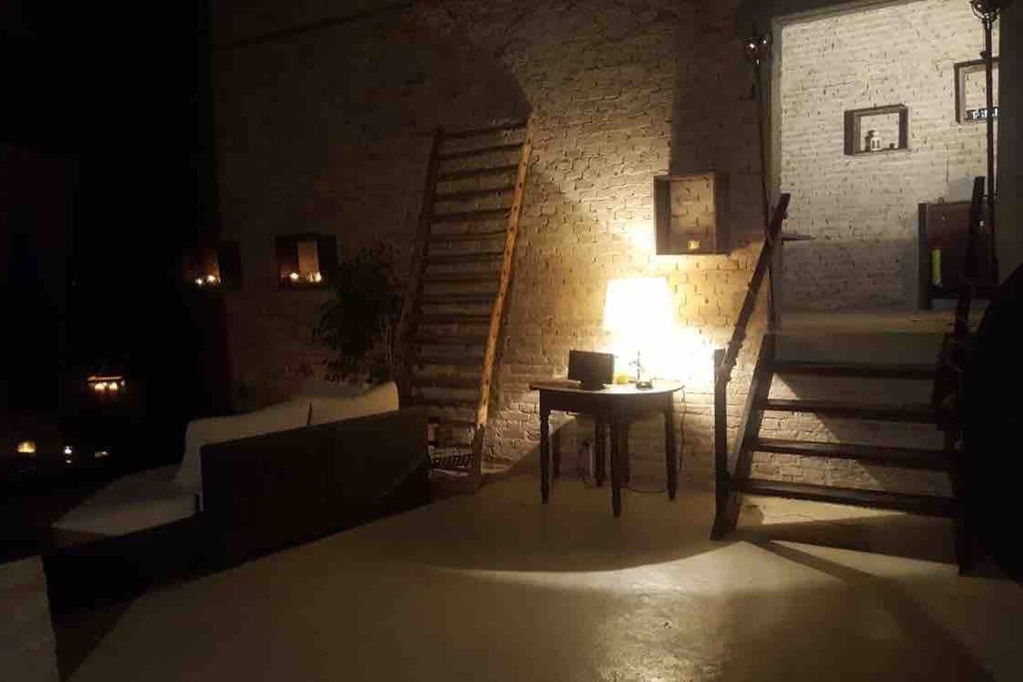 A dimly lit room with a brick wall, a wooden ladder, a small table with a lamp, and a staircase.