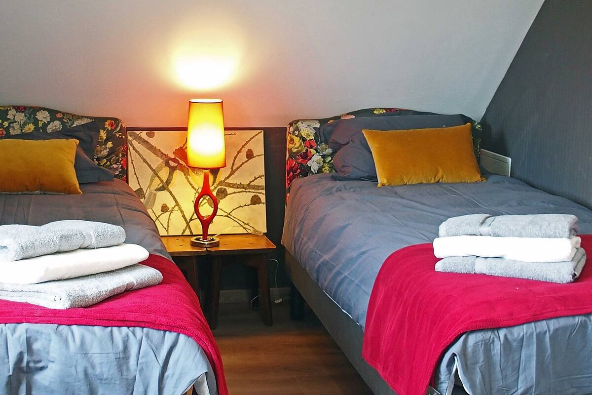 Ein Schlafzimmer mit zwei Betten, jedes mit einer roten Decke und einem weißen Handtuch. Dazwischen steht ein Nachttisch mit einer Lampe.