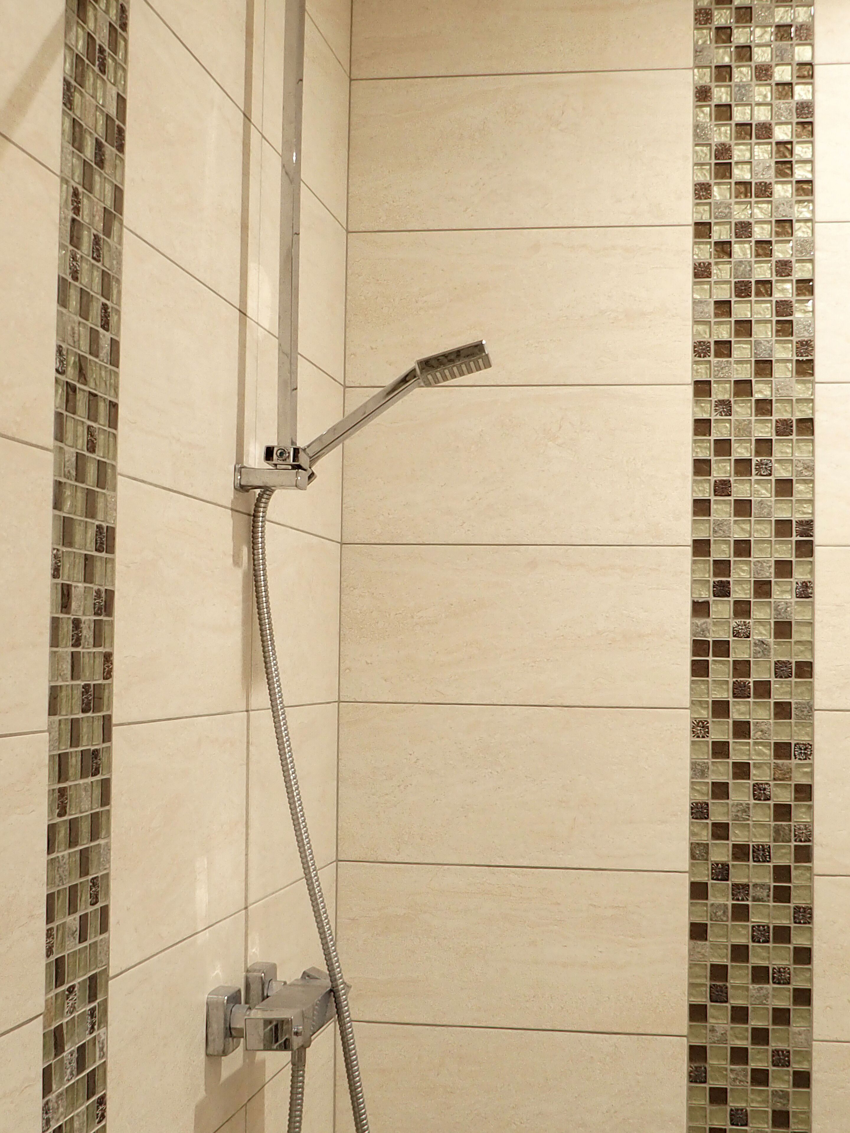 A shower with a handheld showerhead and a flexible hose, set against a tiled wall with a decorative mosaic border.