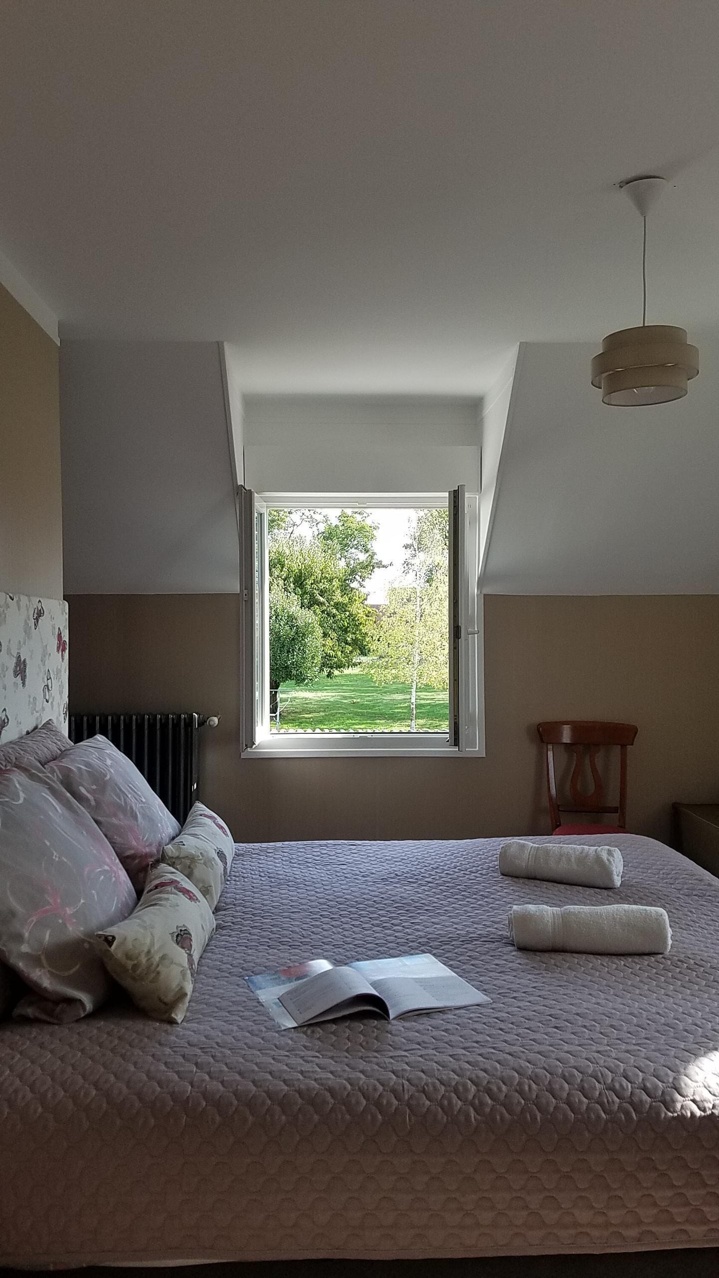 Um quarto com teto inclinado, janela com vista para o verde, cama com travesseiros e um livro, e luminária pendente.