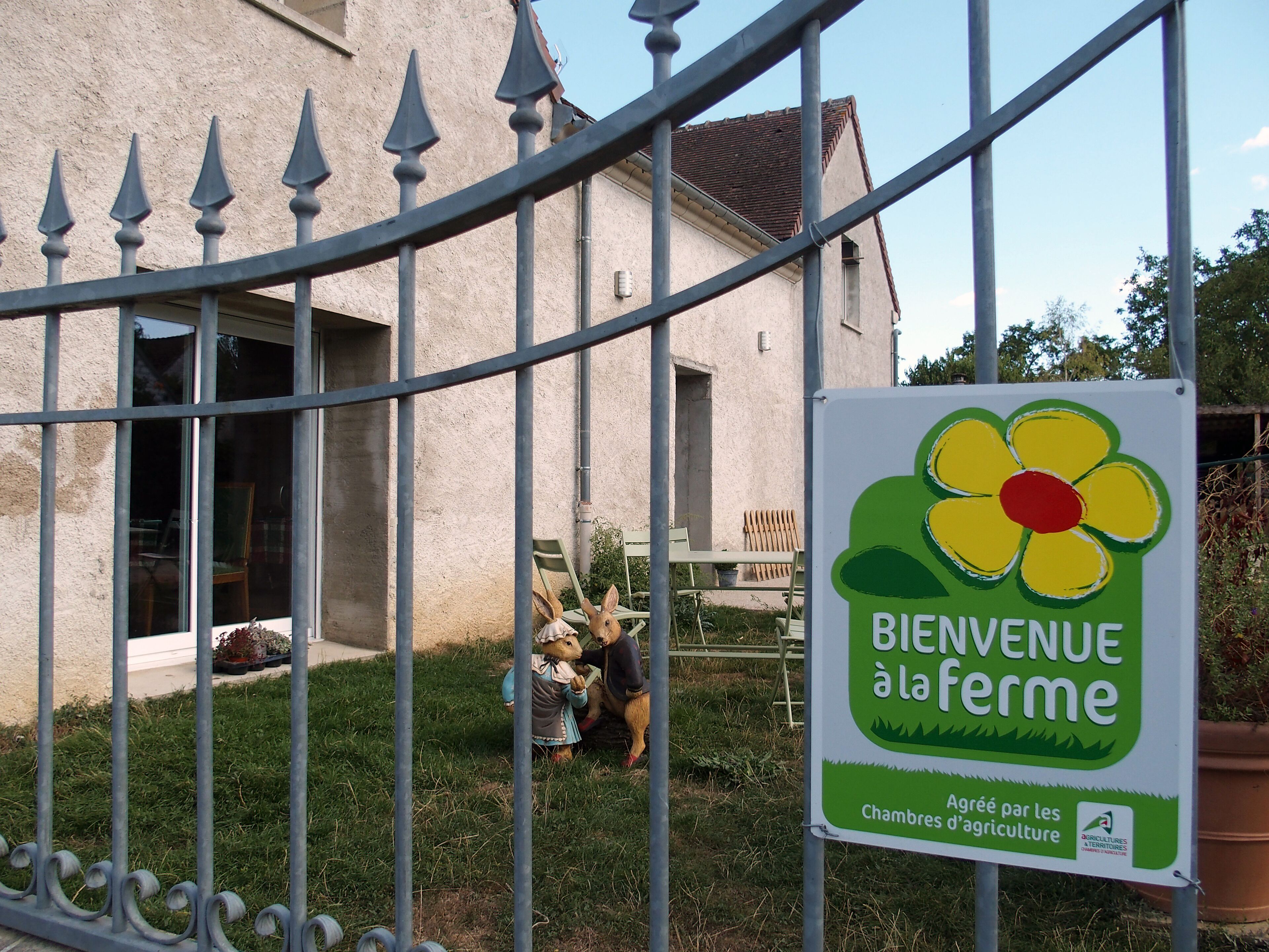 Letrero de bienvenida a visitantes en una granja, con una cerca y un edificio al fondo.