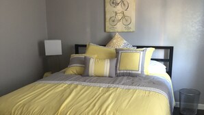 Una cama bien hecha con colcha amarilla y gris, un cabecero negro y almohadones decorativos. Hay una lámpara de pared y un cuadro enmarcado sobre la cama.