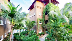 Een binnenplaats met een gebouw, herkenbaar aan het rode dak, meerdere ramen en weelderige begroeiing, waaronder palmbomen en diverse planten.