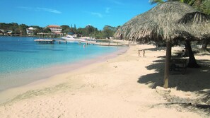 A beach with a thatched hut, clear blue water, and a pier with boats.