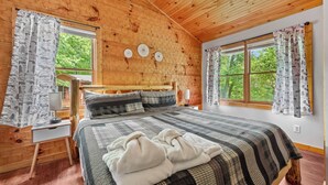 Un dormitorio de cabaña de madera con una cama, mesita de noche y dos ventanas con cortinas.