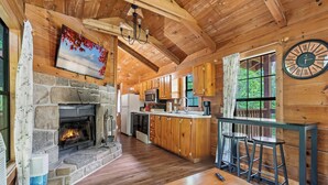 A rustic cabin interior with a stone fireplace, wooden cabinets, a kitchen area, and a bar with stools.
