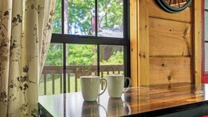 A wooden table with two white mugs in front of a window with a view of greenery.