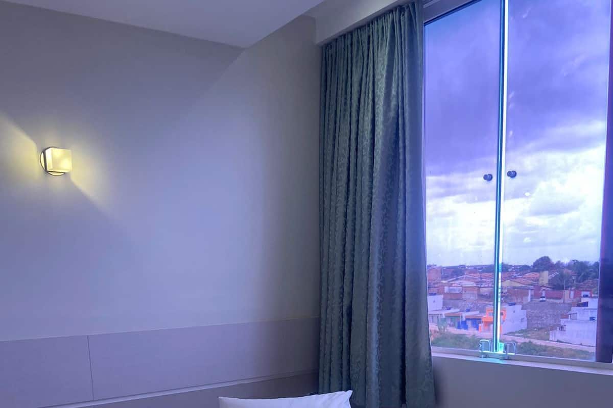 A hotel room with a bed, pillows, a window with curtains, and a view of the cityscape.