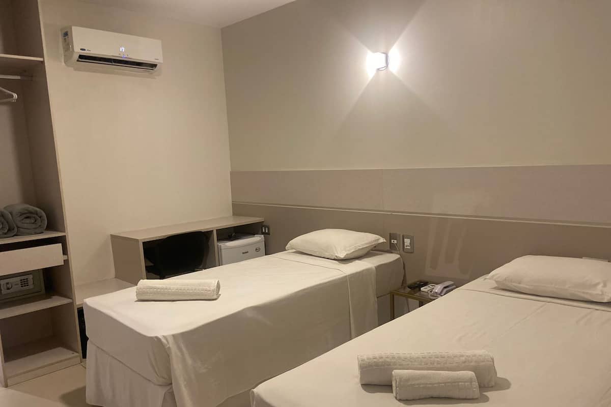 A hotel room with two single beds, a desk, a chair, an air conditioning unit, and a bedside table with a lamp.