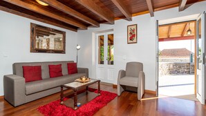 A living room with a grey sofa, red cushions, a wooden coffee table, and a red rug. There is a framed mirror on the wall and a door leading outside. - Raivinha Mother House, A traditional home in the heart of Arco da Calheta (Arco da Calheta, Madeira)