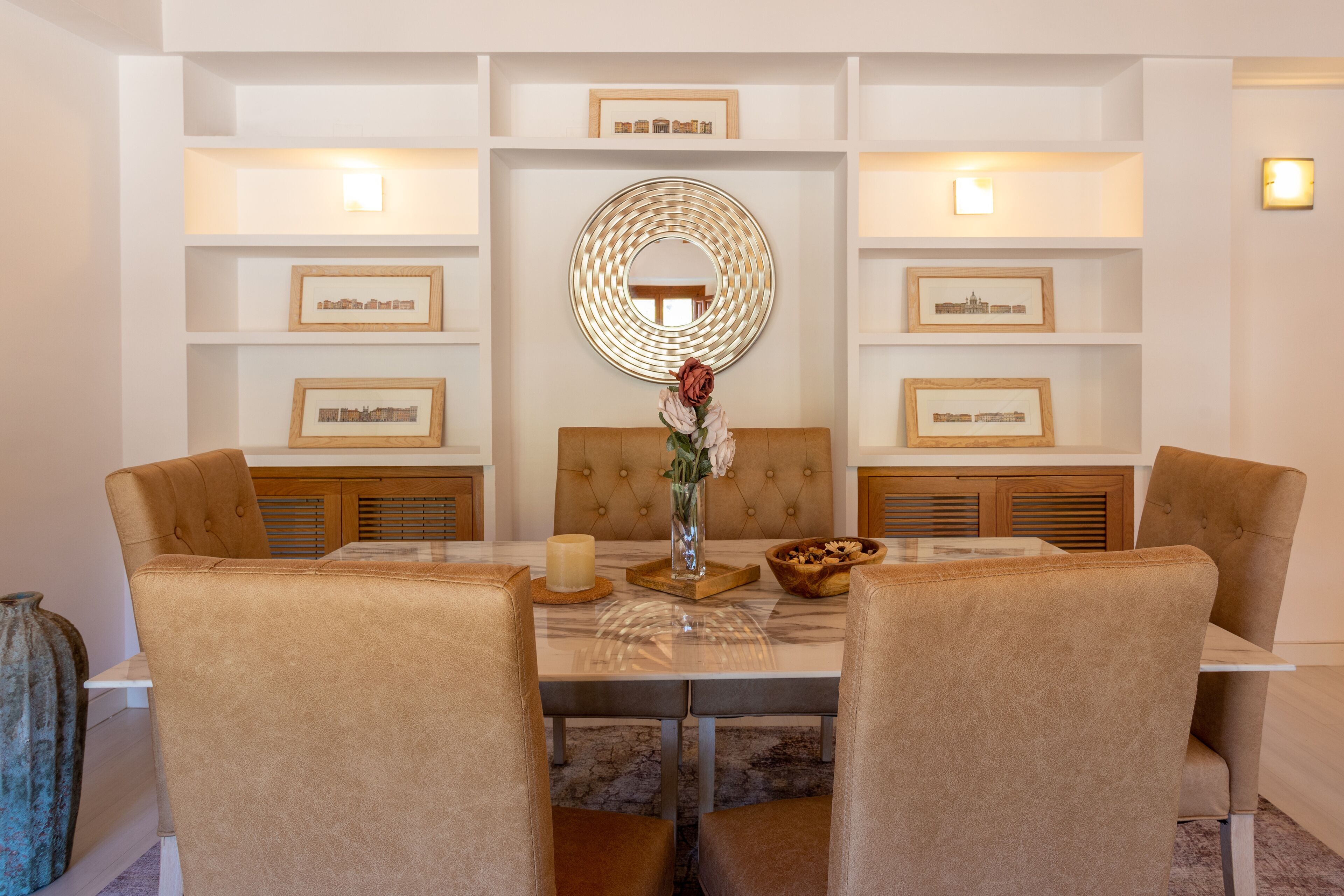 A dining area with a round mirror, framed pictures, and a vase with flowers on the table.