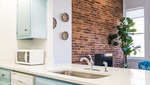 Una cocina con gabinetes azul claro, un microondas blanco, un lavamanos con grifo moderno, una pared decorativa de ladrillo y una planta en maceta.