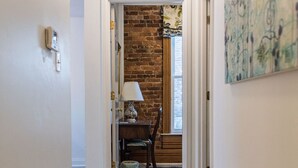 Un pasillo con piso de madera, una puerta que da a una habitación con pared de ladrillo, una mesa con lámpara y un cuadro en la pared.