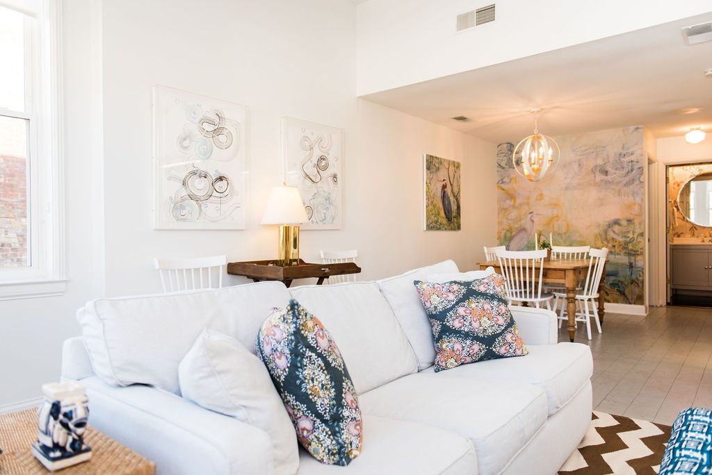 A white sofa with floral cushions, a dining table with chairs, and abstract wall art in a bright room with a large window.