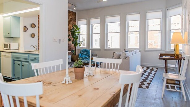 Una cocina moderna con gabinetes color turquesa, un comedor con mesa de madera y sillas blancas, y una zona de estar acogedora con alfombra estampada y una planta en maceta.