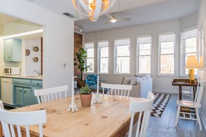 A modern kitchen with teal cabinets, a dining area with a wooden table and white chairs, and a cozy seating area with a patterned rug and a potted plant.