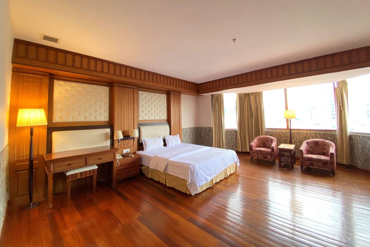 A spacious hotel room with a large bed, a desk with a lamp, two armchairs, and a view of the city through large windows.