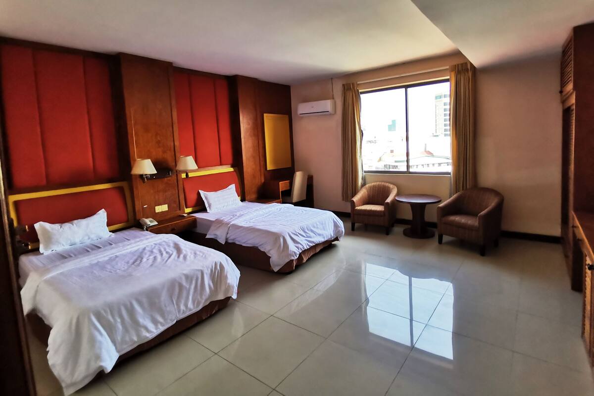 A hotel room with two beds, a small table, a chair, and a window with a view of the city.