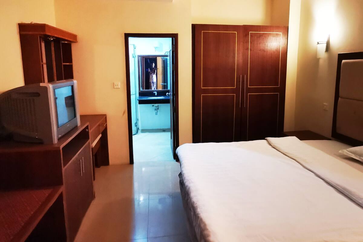 A hotel room with a bed, a TV on a wooden dresser, a wardrobe, and a bathroom visible through an open door.