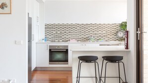 A modern kitchen with white cabinets, a tiled backsplash, a built-in oven, and two black stools. - Beachfront Manly Apartment With Balcony & Parking (Manly)