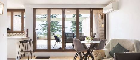 Un vaste espace de vie avec une grande porte-fenêtre coulissante donnant sur une terrasse extérieure. À l’intérieur, on trouve une table à manger avec des chaises, un canapé et une table basse.
