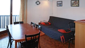 Uma área de refeições com mesa e cadeiras de madeira, um sofá com almofadas vermelhas, um relógio e duas molduras na parede.