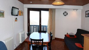 Uma sala de jantar com mesa e cadeiras de madeira, televisão na parede, relógio e vista para uma varanda através de uma porta.