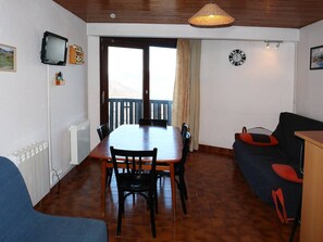 Ein Essbereich mit Holztisch und Stühlen, einem an der Wand befestigten Fernseher, einer Uhr und Blick durch eine Tür auf einen Außenbalkon.