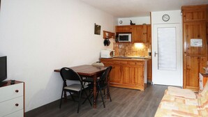 Eine kleine Küche mit Holzschränken, einer Mikrowelle und einem Kühlschrank. Es gibt einen Esstisch mit Stühlen sowie einen Fernseher auf einem Schrank.