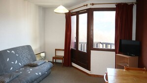 Una habitación pequeña y acogedora con sofá, televisión, una mesa de madera y una ventana con cortinas.