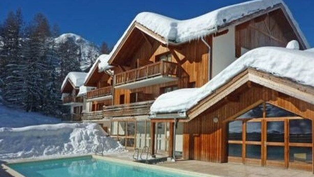 A wooden chalet with a pool, surrounded by snow-covered trees and mountains.