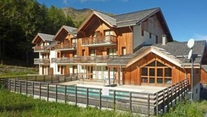 A chalet-style hotel with multiple balconies, a swimming pool, and a mountainous backdrop.
