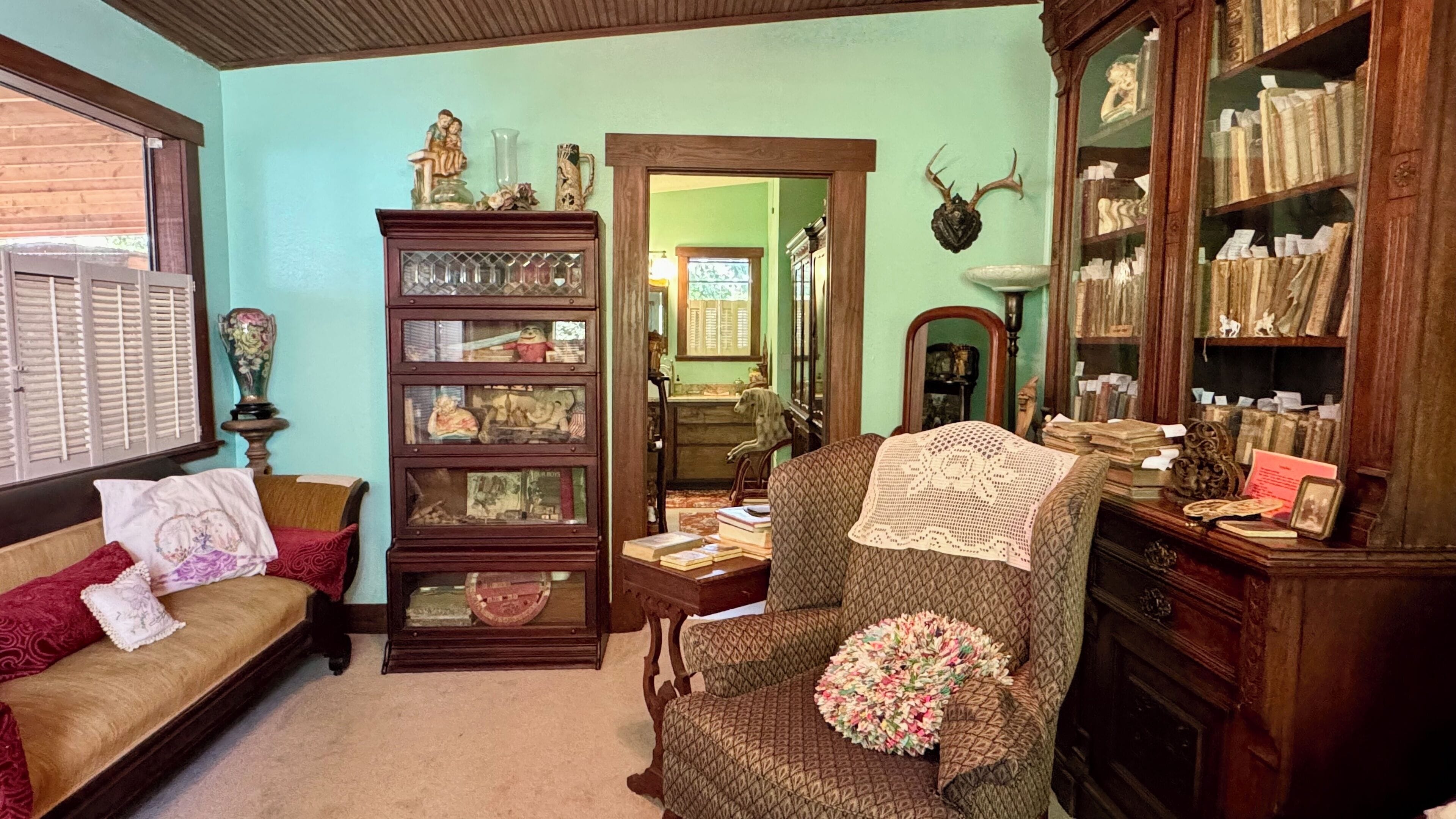 A cozy living room with a wooden sofa, a patterned armchair, a glass-fronted display cabinet, and a bookshelf filled with books and decorative items.