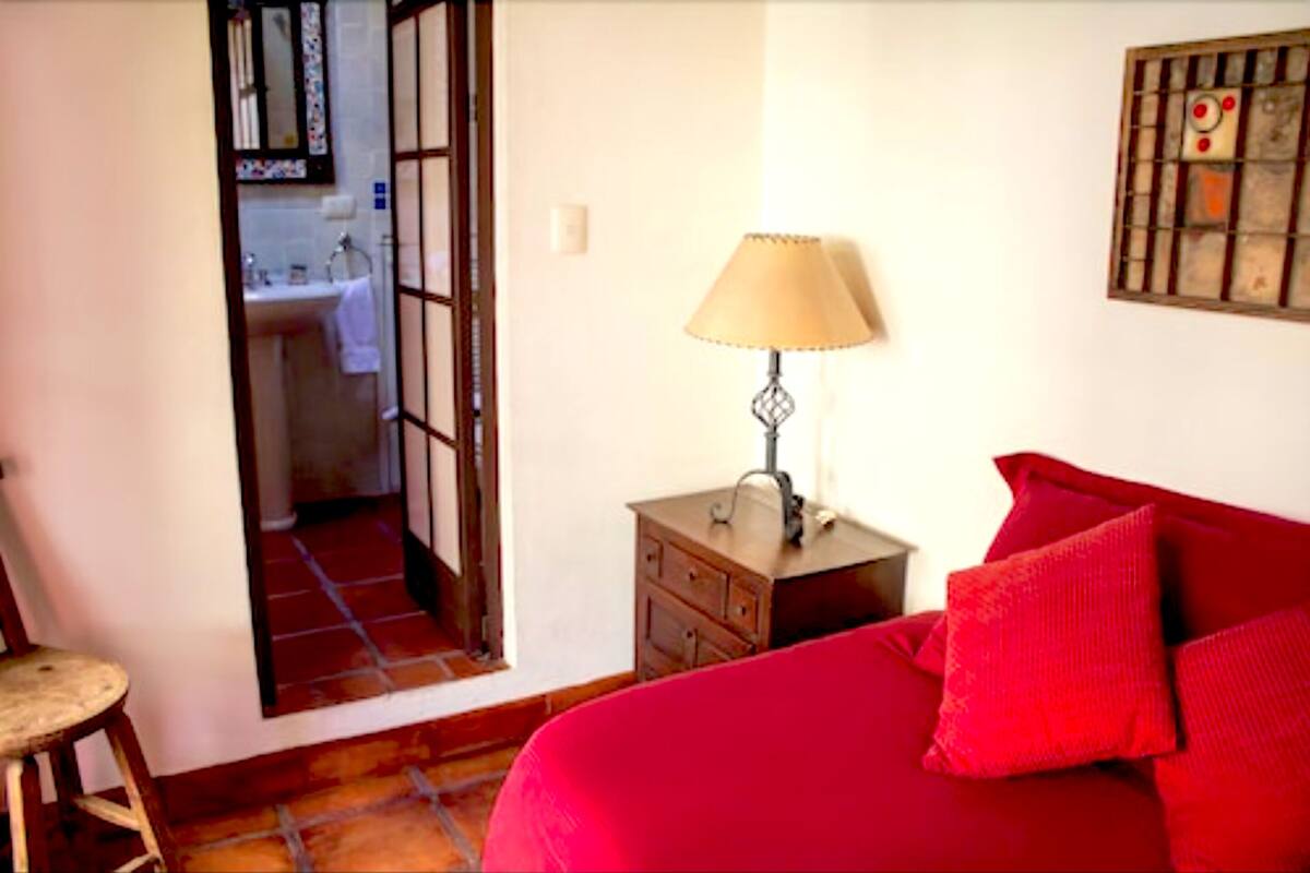 A bedroom with a red bed, a wooden nightstand with a lamp, and a view of a bathroom through an open door.