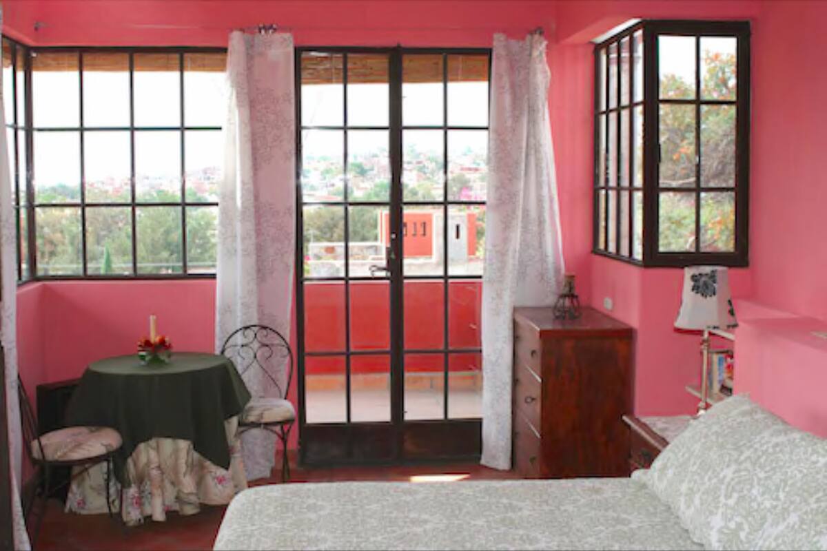 A bedroom with a bed, a small table with a chair, a dresser, and a window with curtains.